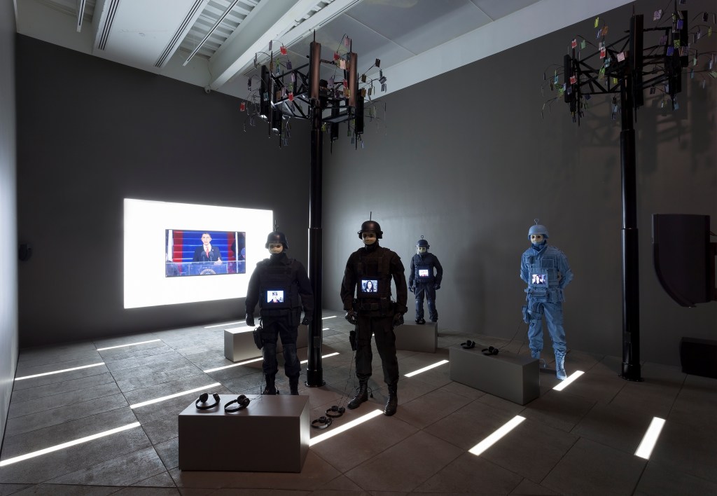 installation view of Josh Kline's "Freedom" exhibition, showing a dark gallery with 4 police officer figures, a video screen in the rear, and 2 black poles rising up from the floor like trees.